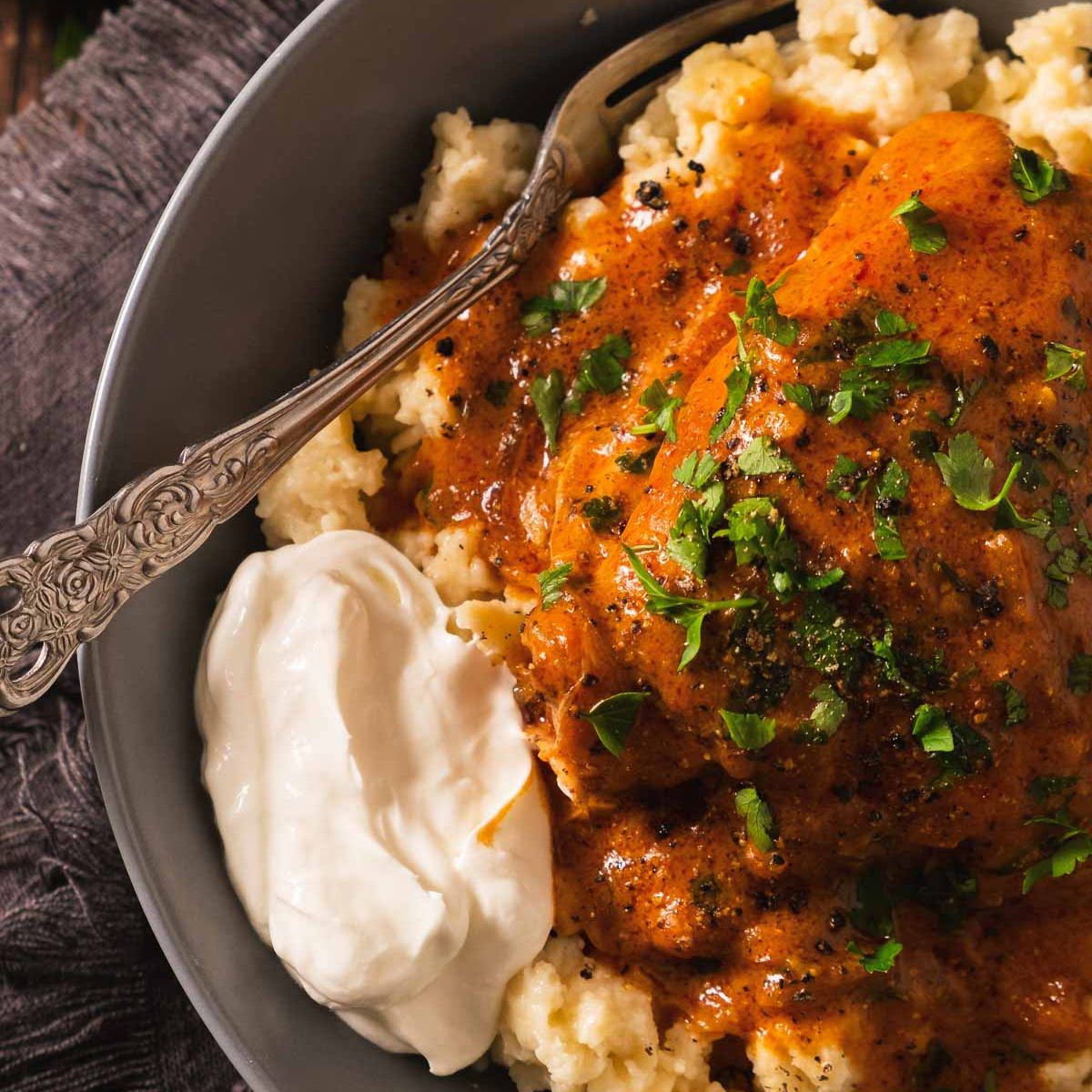 Chicken paprikash on top of spaetzle or nokedli on a plate with a dollop of sour cream on the side.