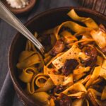 A brown bowl full of haluski, egg noodles with bacon and onion and cabbage. Slices of kielbasa are on top.