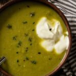 A bowl of creamy green roasted asparagus soup topped with sour cream and fresh thyme leaves.