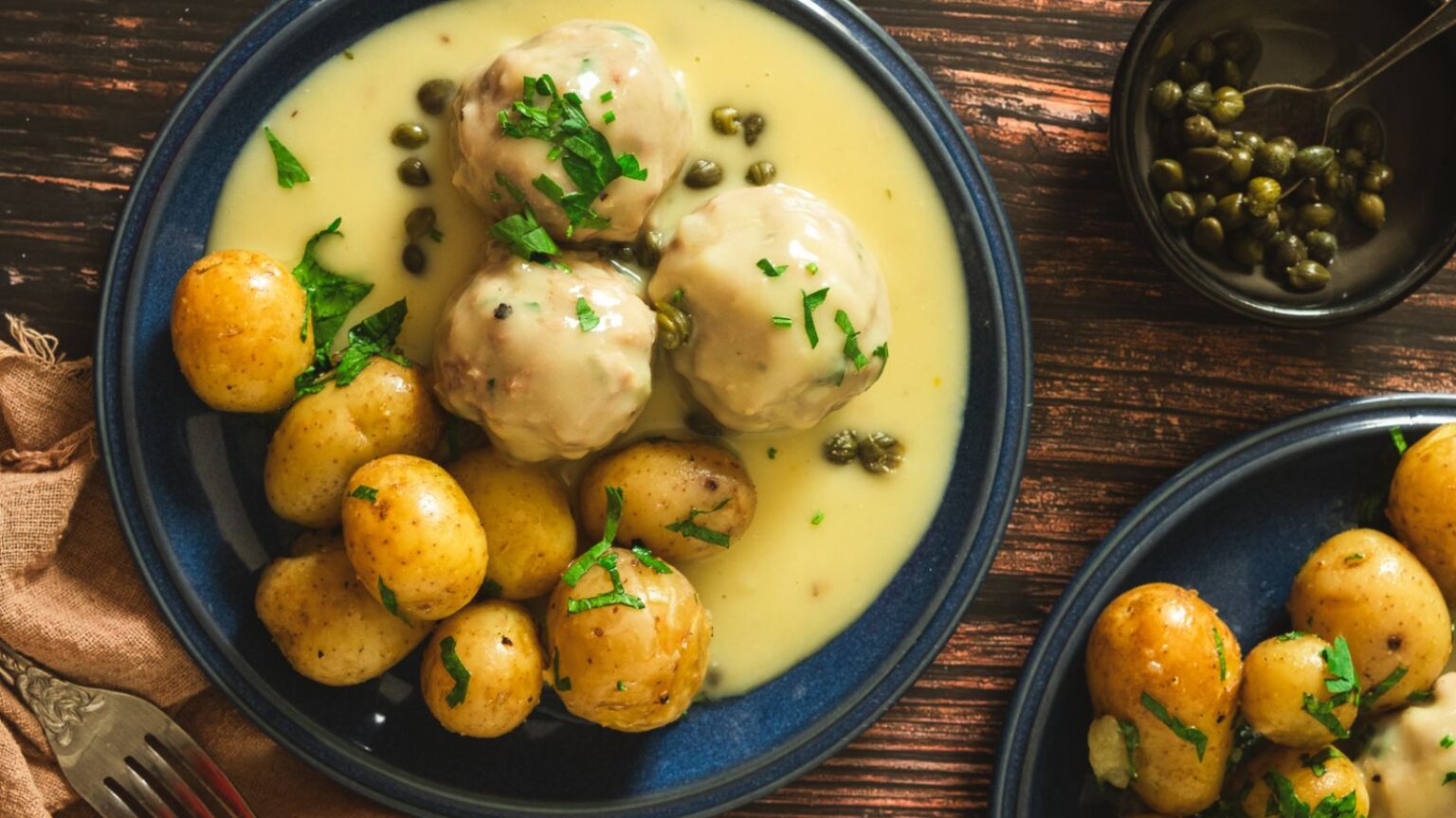 A blue plate of Swedish meatballs covered in creamy sauce, garnished with chopped parsley. The plate includes baby potatoes with parsley on top. To the right, a small bowl contains capers with a spoon. The background features a dark wooden surface.