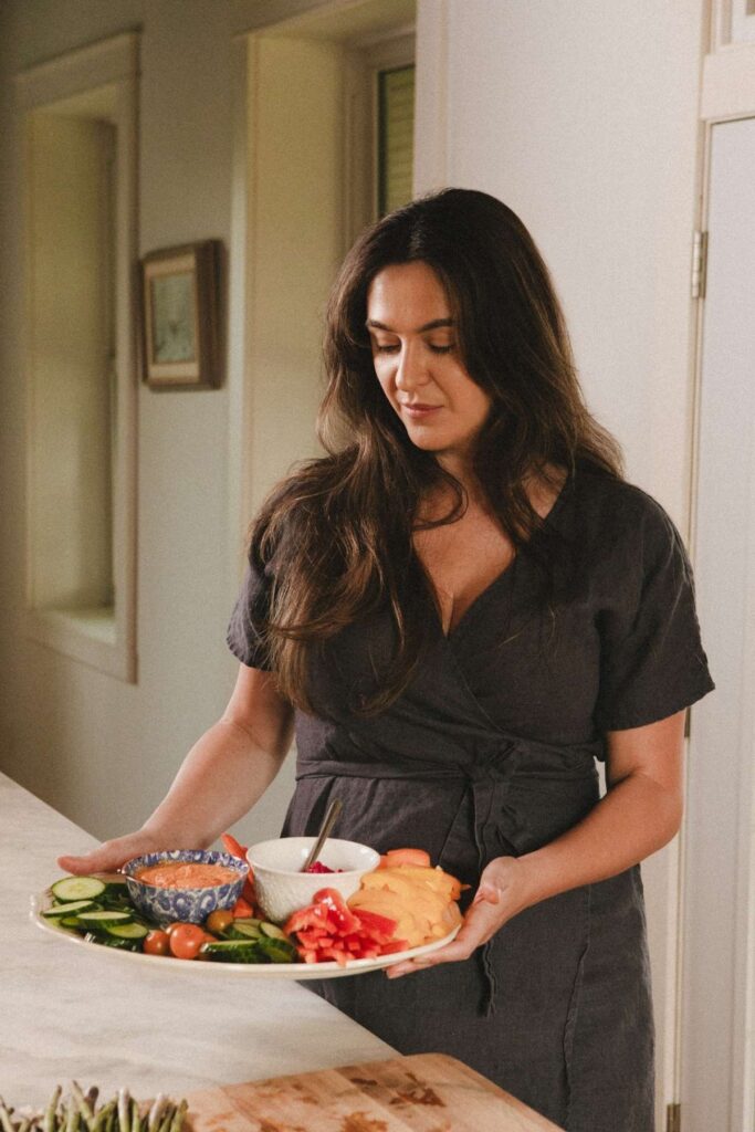 A photo of Jana Dziak, founder of The Peasant's Daughter and Heritage Kitchen Revival standing in her kitchen holding a platter of fresh fruits and veggies with dip.