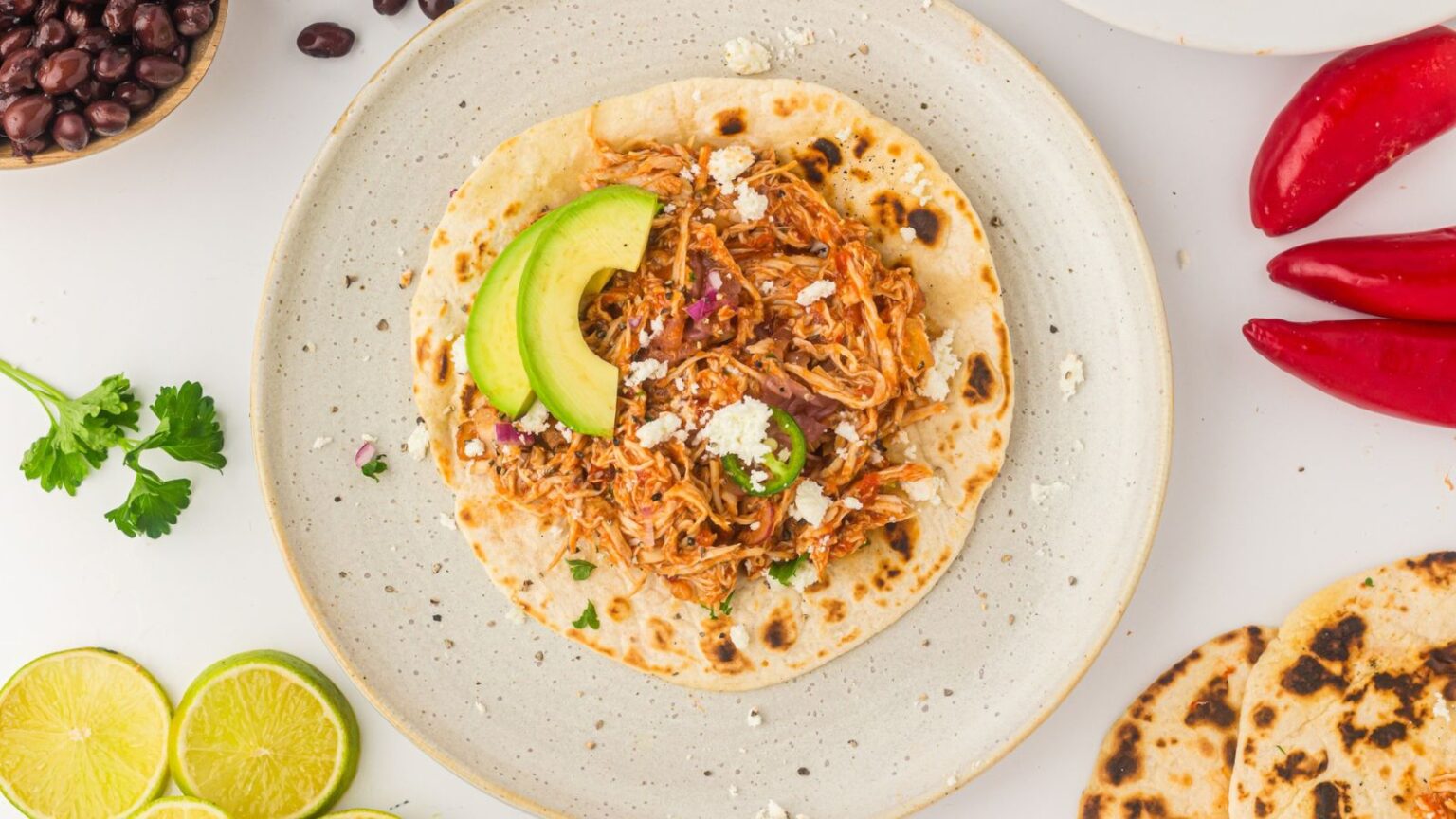 A tortilla loaded with shredded chicken, avocado slices, and crumbled cheese is displayed on a white plate. Surrounding the plate are red chili peppers, grilled tortillas, lime halves, black beans, and fresh cilantro.
