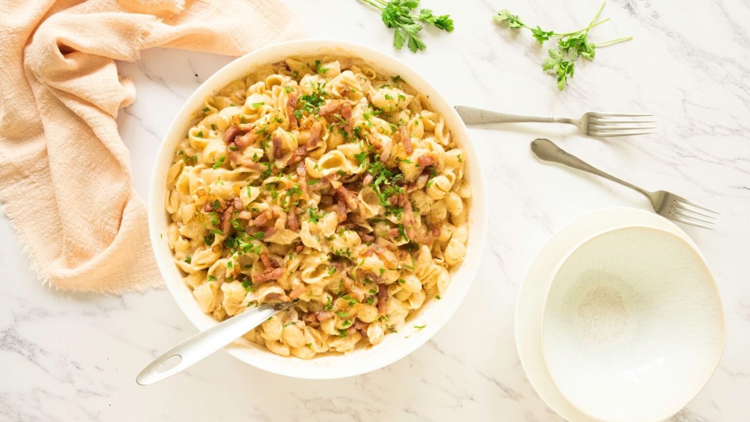 A bowl of pasta is topped with herbs and crispy bacon bits, placed on a marble countertop. A fork rests in the bowl, accompanied by an empty pale green plate and fork beside it. A peach-colored cloth and sprigs of herbs are nearby.