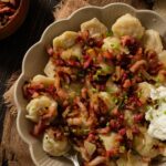 A bowl of pelmeni dumplings smothered in caramelized onions and bacon with sour cream on the side.