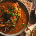 A big bowl of rich, red stew broth with a whole rabbit leg inside. The stew is garnished with fresh parsley.