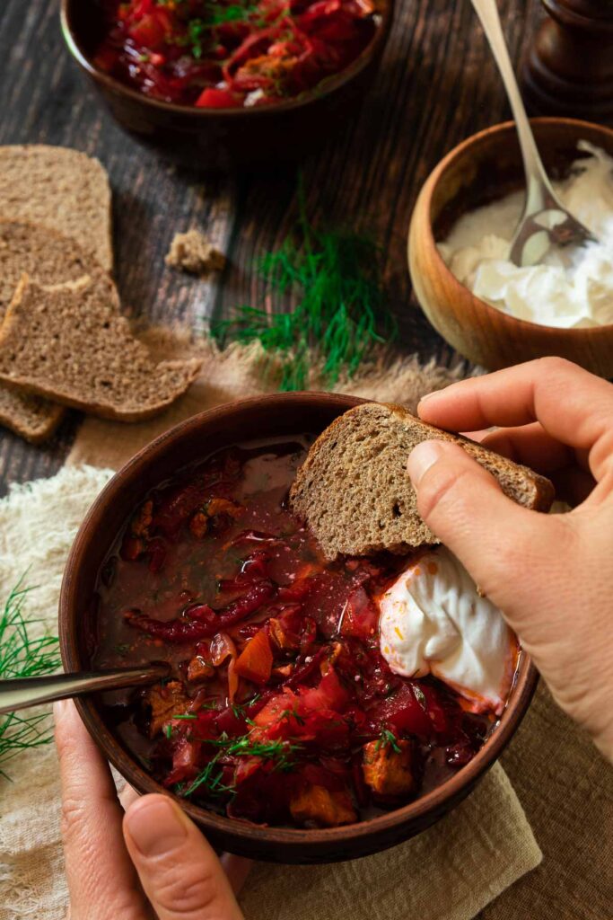 A bowl of traditional Ukrainian borscht beet soup with fresh dill and sour cream. A hand is dipping a piece of dark rye bread into it.