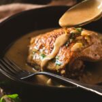 A chicken thigh with golden crispy skin in a thick and creamy deep yellow mustard sauce. A spoon is pouring sauce over the chicken thigh. yellow