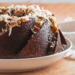 A German chocolate cake made in a bundt pan sitting on a plate with toasted coconut, frosting, ganache, and nuts on top.