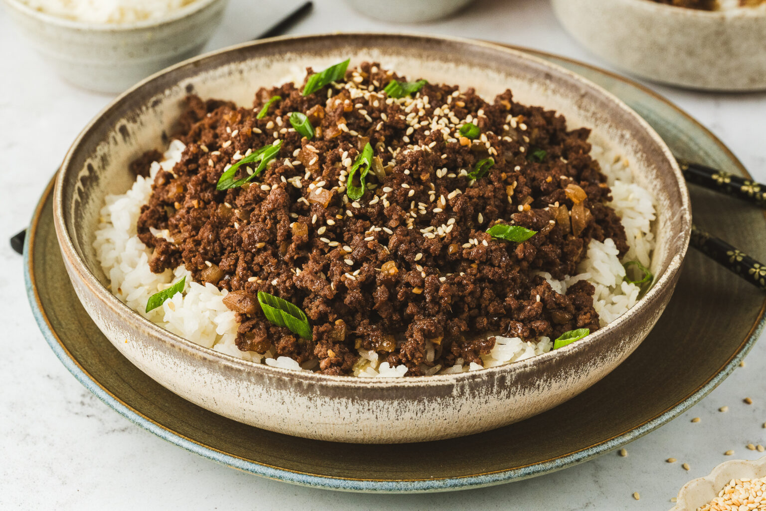 A bowl of white rice topped with flavorful ground beef, garnished with green onions and sesame seeds, on a textured plate.