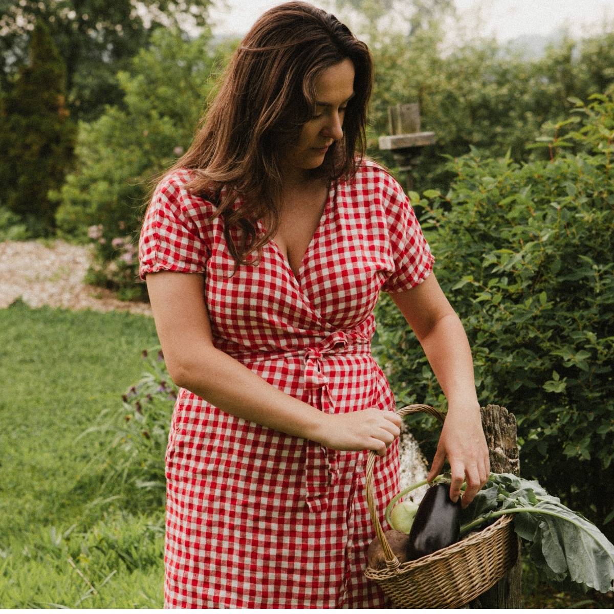 A homemaker in a red gingham dress is outside with a basket of vegetables from her garden.
