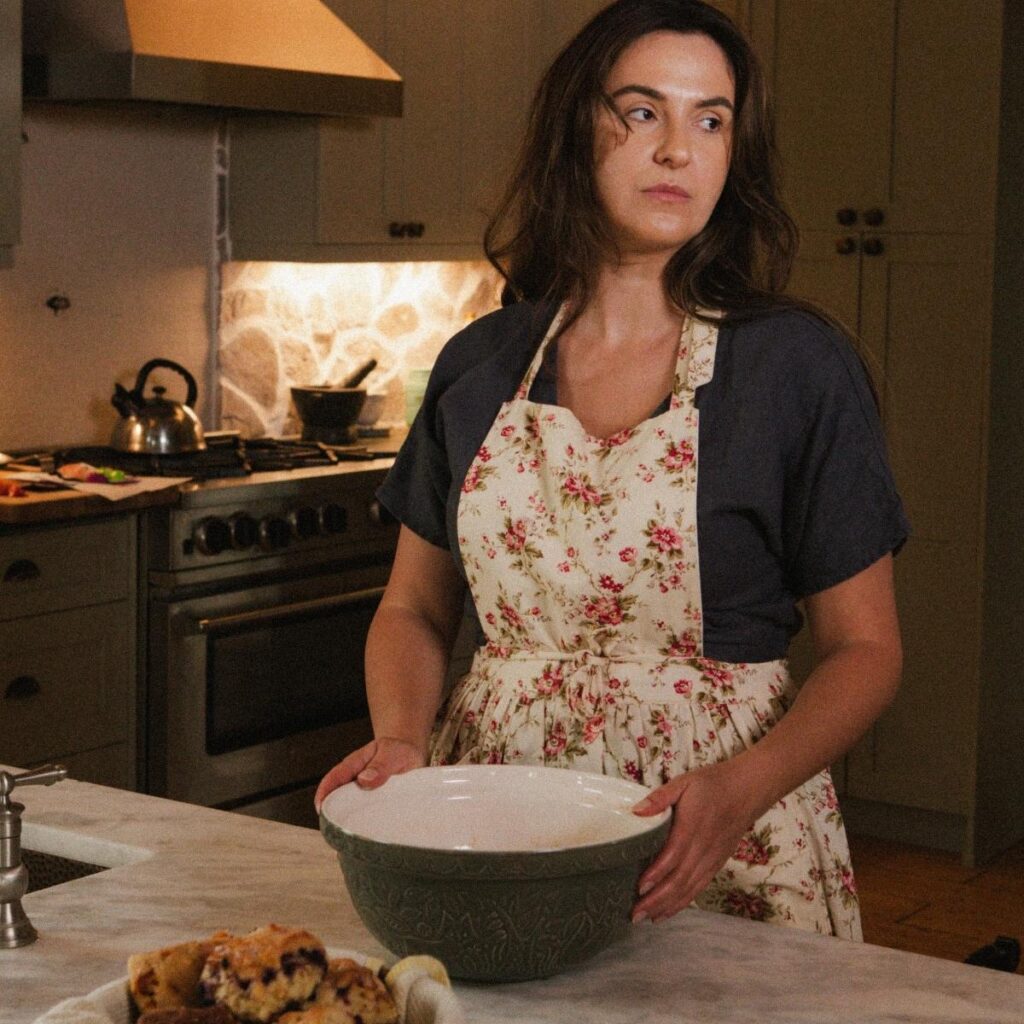A woman stands in her kitchen wearing a dress and apron. She is preparing to bake something.