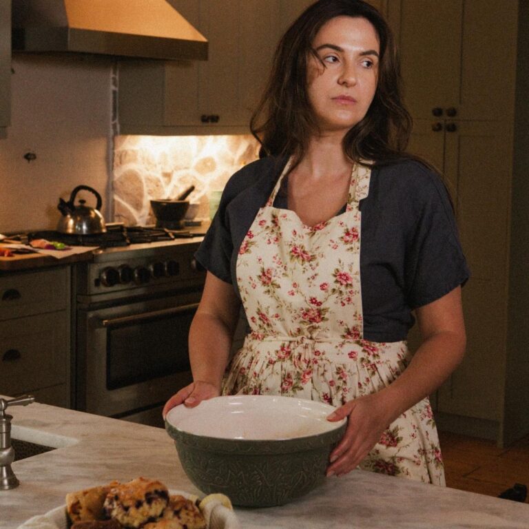 A woman stands in her kitchen wearing a dress and apron. She is preparing to bake something.