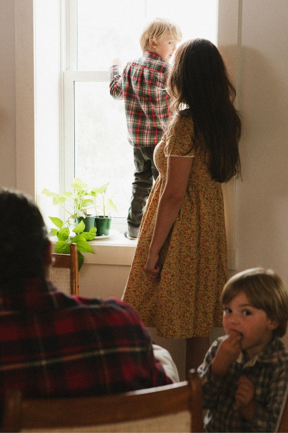 A woman wearing a bright, floral dress stands at the window with her son while her other son and husband sit at the dining room table.