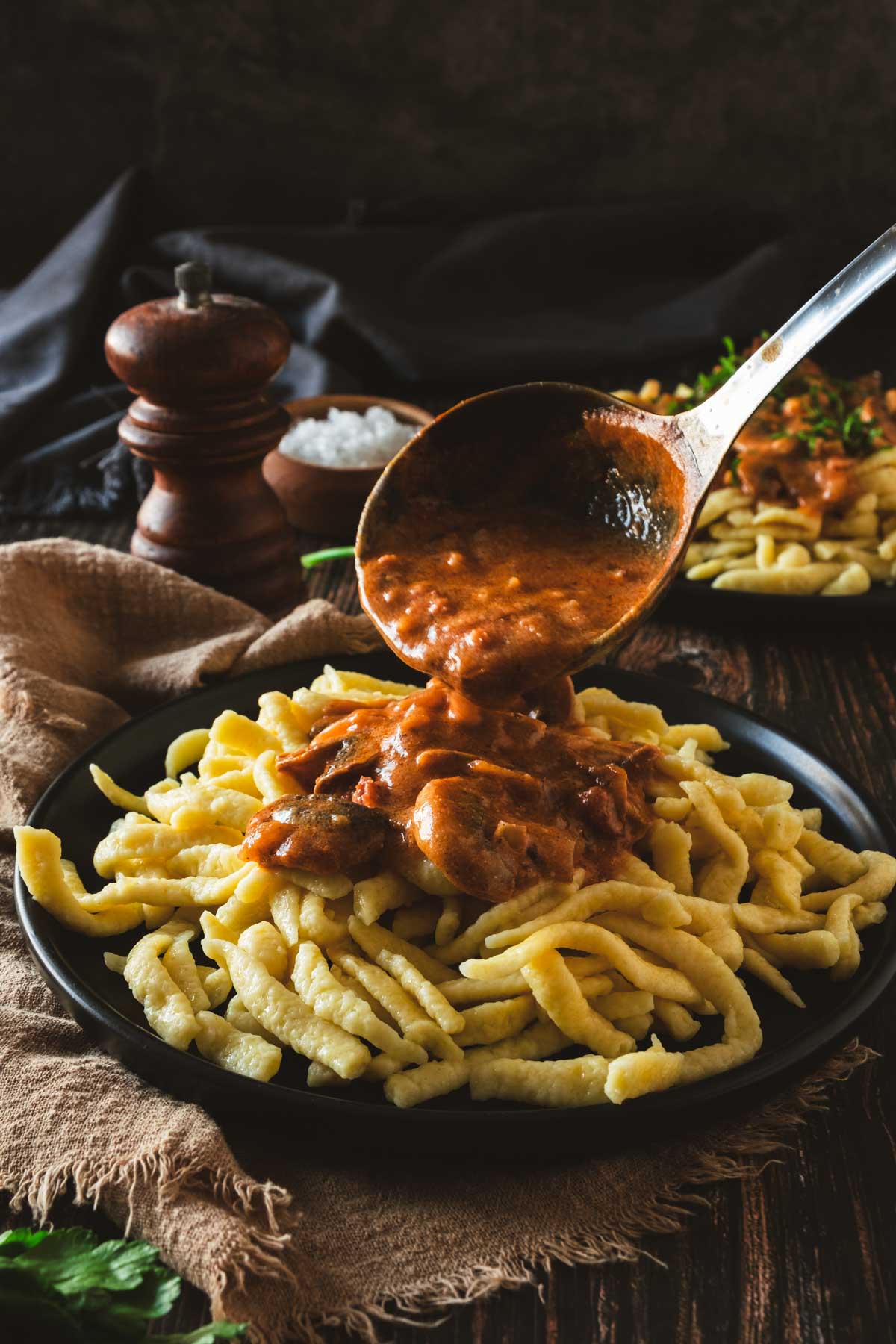 A ladle of creamy mushroom gravy is being poured over spaetzle noodle dumplings.