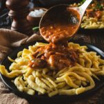 A ladle of creamy mushroom gravy is being poured over spaetzle noodle dumplings.