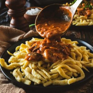 A ladle of creamy mushroom gravy is being poured over spaetzle noodle dumplings.