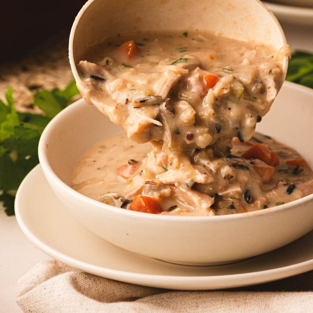 Creamy chicken and wild rice soup being ladled into a bowl, with pieces of chicken, wild rice, and vegetables in a rich, creamy broth.
